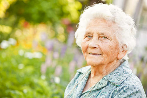 An elderly woman with white hair and a green shirt smiles as she sits outside in the sun amid colorful flowers.